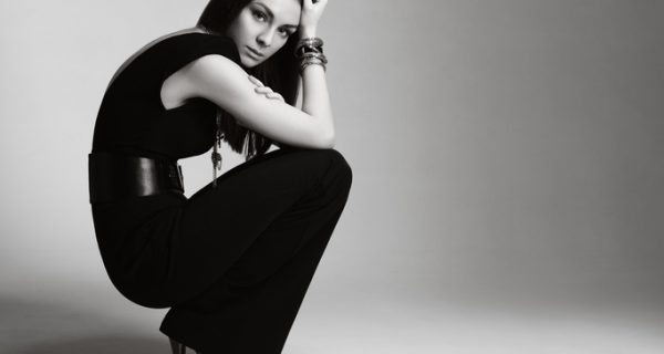 Fashionable young woman wearing black jumpsuit and accessories and sitting in studio.